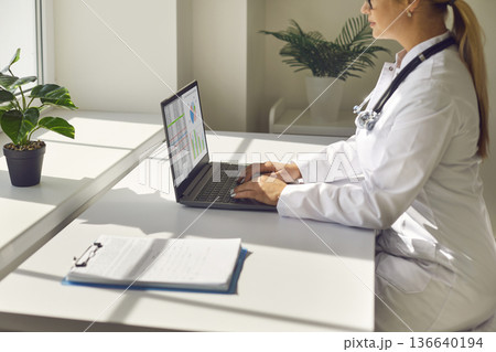 Female doctor working online on laptop in bright medical office, telehealth concept 136640194