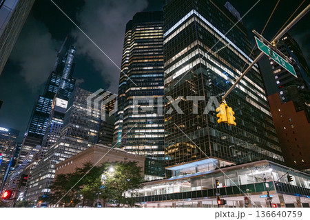 Tall glass skyscrapers tower above East 53rd Street in New York City, illuminated by office lights and traffic signals. A yellow streetlight and dark sky add contrast to the vibrant urban night scene. Tall glass skyscrapers tower above East 53rd Street in New York City, illuminated by office lights and traffic signals. A yellow streetlight and dark sky add contrast to the vibrant urban night scene. 136640759