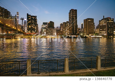 The New York City skyline shines with illuminated skyscrapers beside the Queensboro Bridge at twilight. The river reflects golden lights, and a quiet metal fence lines the calm waterfront in the 136640770