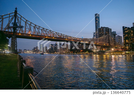 The Queensboro Bridge glows with lights as it stretches over the East River, connecting to Manhattan's vibrant skyline. The illuminated buildings and bridge reflect beautifully on the calm evening The Queensboro Bridge glows with lights as it stretches over the East River, connecting to Manhattan's vibrant skyline. The illuminated buildings and bridge reflect beautifully on the calm evening 136640775