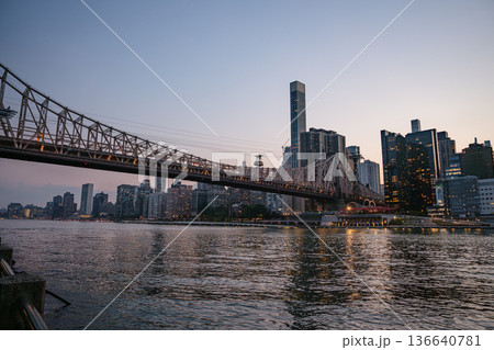 The Queensboro Bridge spans across the East River as Manhattan's illuminated skyline reflects in the calm water. The scene captures the peaceful beauty of the city during the last light of day. 136640781