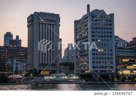 Two uniquely shaped concrete and glass office towers rise above the East River in Manhattan. The buildings are illuminated with warm lights as evening falls over the city. 136640787