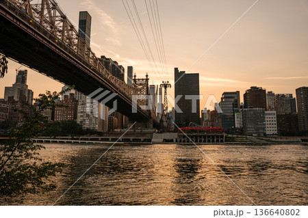 The Queensboro Bridge stretches over the East River at sunset with Manhattan skyscrapers in the background. The water reflects the golden sky, creating a peaceful urban scene. The Queensboro Bridge stretches over the East River at sunset with Manhattan skyscrapers in the background. The water reflects the golden sky, creating a peaceful urban scene. 136640802