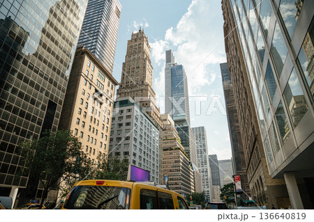 A yellow taxi drives down a bustling street in Midtown Manhattan. Historic and modern skyscrapers rise into a bright summer sky. A yellow taxi drives down a bustling street in Midtown Manhattan. Historic and modern skyscrapers rise into a bright summer sky. 136640819