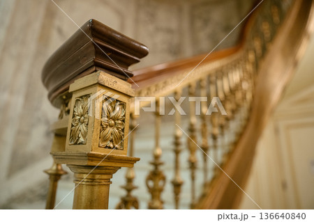 A close-up view of a staircase railing shows a golden floral carving and a dark polished wooden handrail. The blurred background reveals elegant balusters and a softly lit vintage interior. A close-up view of a staircase railing shows a golden floral carving and a dark polished wooden handrail. The blurred background reveals elegant balusters and a softly lit vintage interior. 136640840