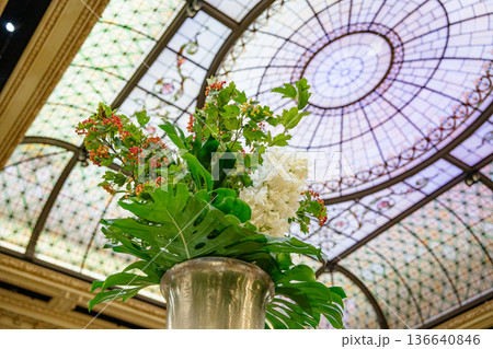 A luxurious flower arrangement stands in a tall silver vase beneath a stained glass dome ceiling. Bright green leaves, white blossoms, and red berries add vivid color to the ornate indoor setting. A luxurious flower arrangement stands in a tall silver vase beneath a stained glass dome ceiling. Bright green leaves, white blossoms, and red berries add vivid color to the ornate indoor setting. 136640846