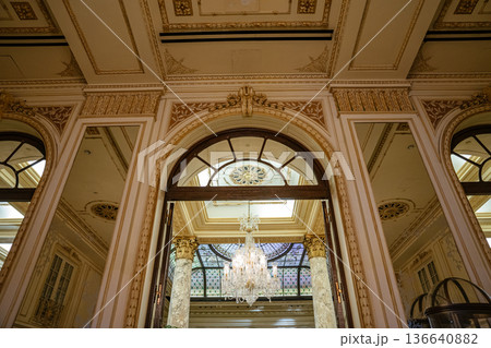 An ornate arched doorway opens to a grand chandelier and stained glass ceiling framed by marble columns. Detailed moldings, gold accents, and architectural symmetry reflect classic luxury in a 136640882