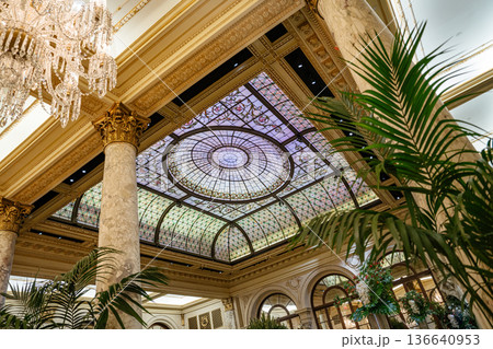 Beautiful stained glass dome ceiling above an opulent hotel lobby with marble columns and lush indoor plants. Classic architecture and intricate details create an atmosphere of elegance and grandeur. Beautiful stained glass dome ceiling above an opulent hotel lobby with marble columns and lush indoor plants. Classic architecture and intricate details create an atmosphere of elegance and grandeur. 136640953