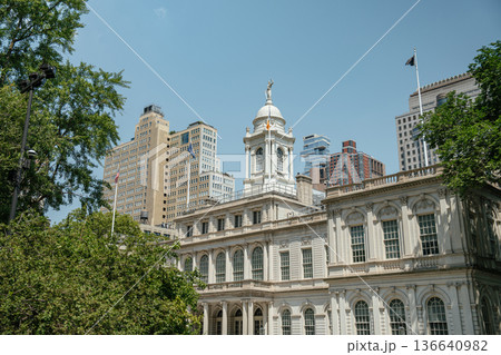 Detailed view of the historic New York City Hall with its iconic white clock tower. Surrounding trees and modern skyscrapers highlight the contrast of old and new architecture. 136640982