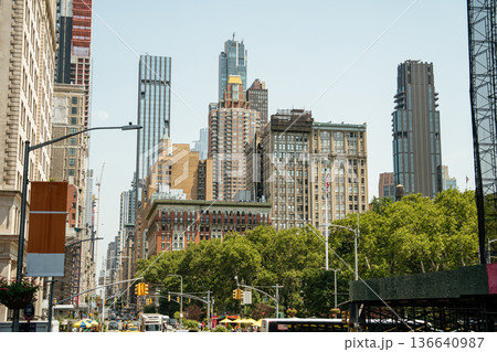 A view of Midtown Manhattan shows a mix of historic and modern skyscrapers towering above green trees. The busy street below is filled with yellow taxis, buses, and city life on a sunny day. A view of Midtown Manhattan shows a mix of historic and modern skyscrapers towering above green trees. The busy street below is filled with yellow taxis, buses, and city life on a sunny day. 136640987