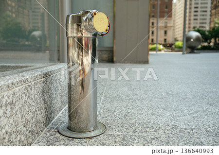 A shiny stainless steel fire department connection stands mounted on a granite sidewalk near a glass building. Urban architecture and blurred buildings appear in the background on a sunny day. 136640993