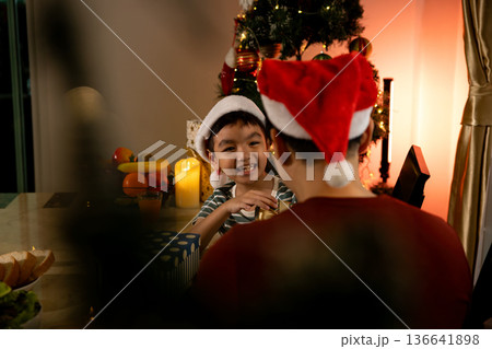 Father and son wearing Santa hats celebrating Christmas at home, smiling at dinner table with decorated tree, wrapped gift and candlelight, warm family holiday tradition, festive evening 136641898