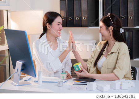 Two asian businesswoman team high five celebrate success cosmetic packaging design project. Two asian businesswoman team high five celebrate success cosmetic packaging design project. 136642583