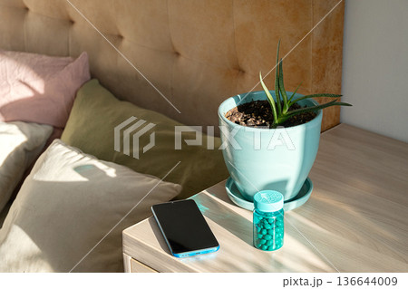 Smartphone, potted plant, and bottle of pills on nightstand beside bed. Smartphone, potted plant, and bottle of pills on nightstand beside bed. 136644009