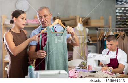 Girl customer examines finished product in presence of mature man tailor, guy sew in background 136644448