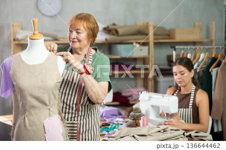 Elderly dressmaker stands near a mannequin and checks the dress against the background of the assistant 136644462
