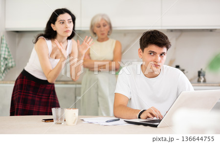 Young guy while working on laptop listens to claims of elderly mother and young sister girl Young guy while working on laptop listens to claims of elderly mother and young sister girl 136644755