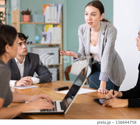 Confident young businesswoman standing near table where sitting colleagues talking and discussing ideas in office Confident young businesswoman standing near table where sitting colleagues talking and discussing ideas in office 136644756