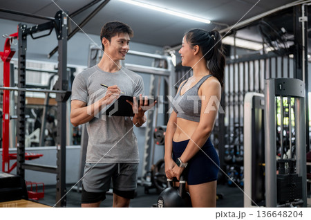 Fitness trainer coach man holding exercise plan clipboard and talking to asian woman client in gym Fitness trainer coach man holding exercise plan clipboard and talking to asian woman client in gym 136648204