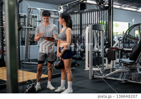 Fitness trainer coach man holding clipboard talking about exercise plan with a woman client in gym. 136648207