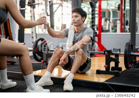 Asian sportswear man sitting on gym floor holding water clinking bottles with woman friend on bench. Asian sportswear man sitting on gym floor holding water clinking bottles with woman friend on bench. 136648242