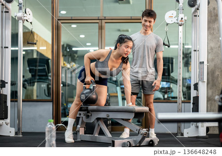 Happy fitness trainer coach man teaching asian woman client holding kettlebell exercising in the gym 136648248
