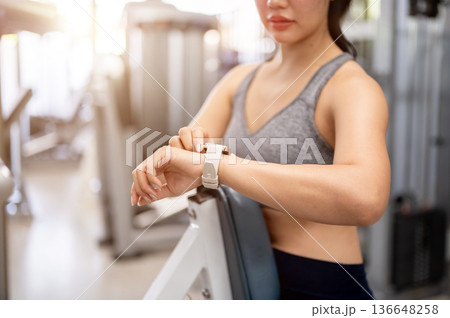 Close up of asian woman in sportswear standing looking at time on wristwatch after exercising in gym Close up of asian woman in sportswear standing looking at time on wristwatch after exercising in gym 136648258