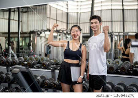 Pretty asian woman fitness trainer holding laptop raising a fist with asian man client in the gym. 136648270
