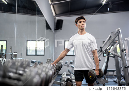 Asian man fitness trainer coach or a client standing and holding a dumbbell while exercising in gym. 136648276