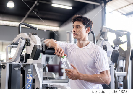 Asian man client or fitness trainer coach holding water bottle taking a rest after exercising in gym 136648283