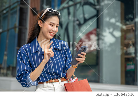 Pretty asian woman holding and looking at phone carrying shopping bags while walking outside a mall. 136648510