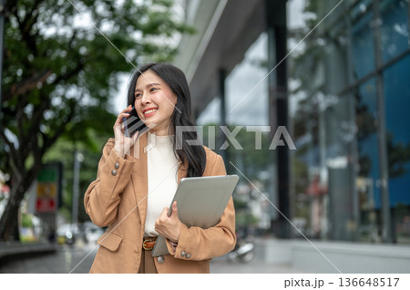 Pretty asian woman office worker talking on phone and carrying laptop walking outside shopping mall. Pretty asian woman office worker talking on phone and carrying laptop walking outside shopping mall. 136648517