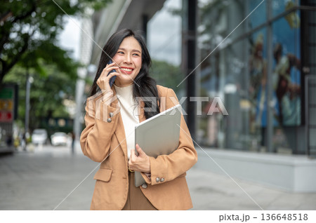 Pretty asian woman office worker talking on phone as carrying laptop walking outside a shopping mall 136648518