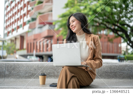 Pretty asian woman office worker working on laptop while sitting on stone bench in a garden or park. 136648521
