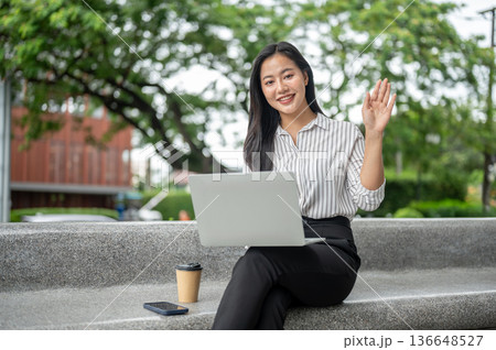 Pretty asian woman office worker waving hand over laptop and coffee sitting on stone bench in garden Pretty asian woman office worker waving hand over laptop and coffee sitting on stone bench in garden 136648527