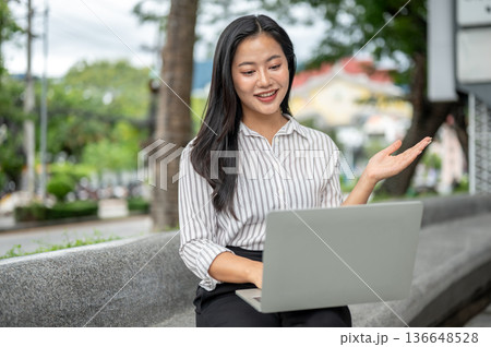 Pretty asian woman office worker explaining with hand over laptop sitting on stone bench in a garden 136648528