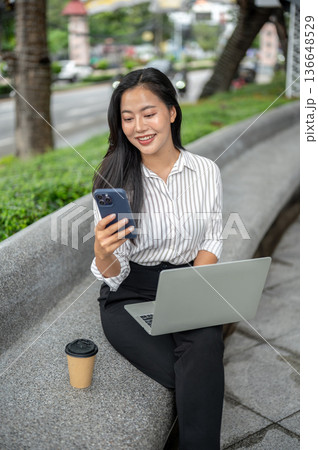 Pretty asian woman office worker looking at phone over laptop and sitting on stone bench in a garden Pretty asian woman office worker looking at phone over laptop and sitting on stone bench in a garden 136648529