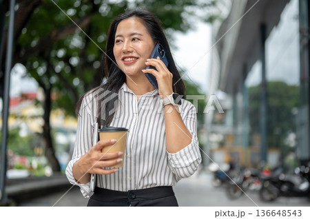 Asian woman office worker holding coffee cup as talking on phone while walking outside shopping mall 136648543