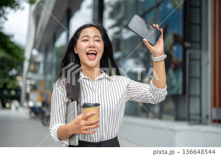 Asian woman office worker holding coffee waving hand with phone while walking outside shopping mall. 136648544