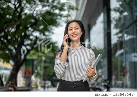 Pretty woman office worker holding talking on phone carrying laptop as walking outside shopping mall Pretty woman office worker holding talking on phone carrying laptop as walking outside shopping mall 136648545