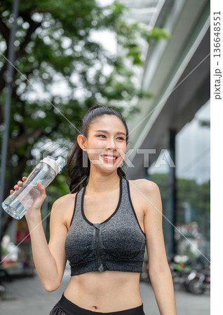 Asian woman in sportswear holding water bottle standing taking a rest after exercising aside a mall. 136648551