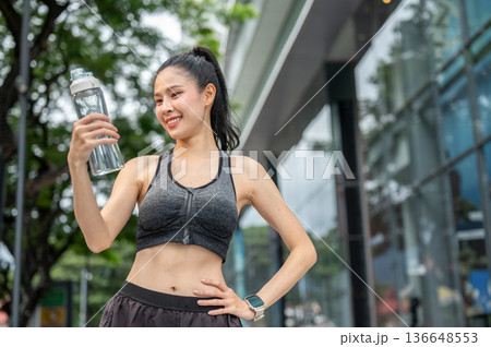 Asian woman in sportswear holding looking at water bottle with hand on waist resting after exercise. 136648553