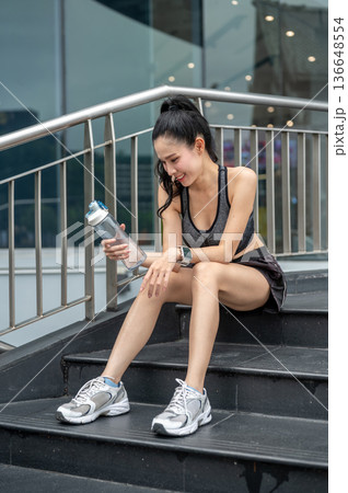 Asian woman in sportswear holding looking water bottle while sitting on stair resting after exercise 136648554