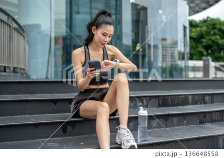 Asian sportswear woman holding phone looking at time watch sitting on stairs resting after exercise. Asian sportswear woman holding phone looking at time watch sitting on stairs resting after exercise. 136648558