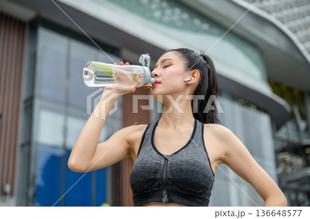 Asian woman in sportswear with earbud closing eyes drinking water after running exercise aside mall. 136648577