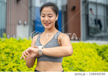 Asian woman in sportswear listening to music looking at smartwatch while running exercising in park. 136648580