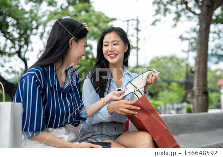 Asian woman looking at cloth in a friend's shopping bag while sitting on bench at mall's garden park 136648592