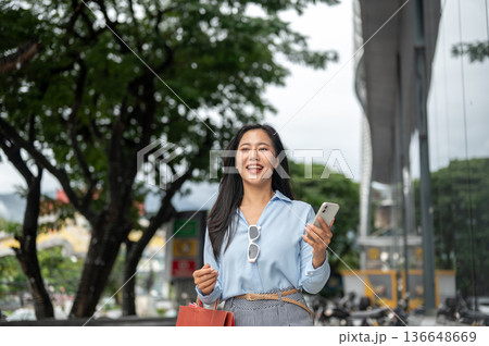 Pretty asian woman carrying shopping bags and holding phone standing walking outside a shopping mall Pretty asian woman carrying shopping bags and holding phone standing walking outside a shopping mall 136648669