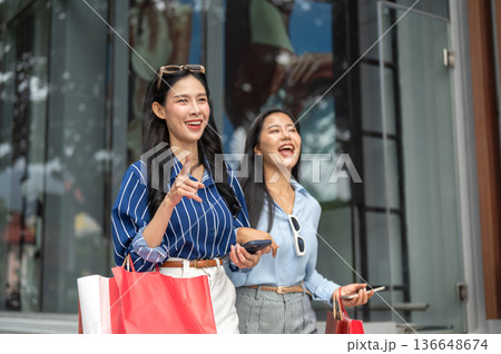 Pretty asian woman and laughing friend carrying shopping bag walking together outside shopping mall. Pretty asian woman and laughing friend carrying shopping bag walking together outside shopping mall. 136648674