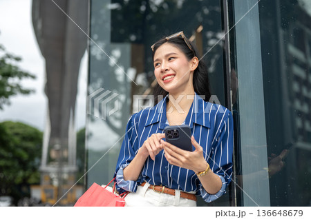 Pretty asian woman carrying shopping bag and holding looking at phone while standing outside a mall. Pretty asian woman carrying shopping bag and holding looking at phone while standing outside a mall. 136648679